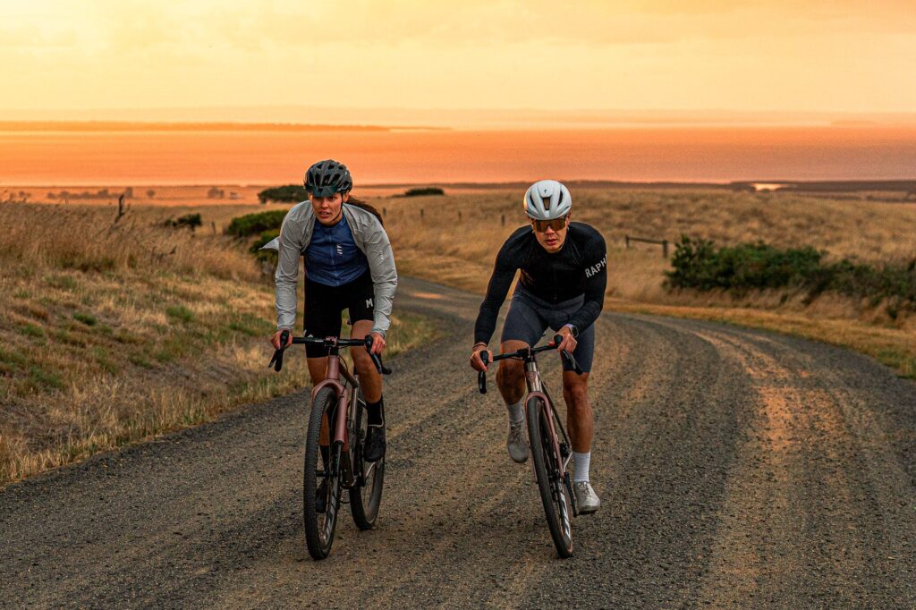 Two riders on titanium gravel bikes climbing a gravel road at sunset