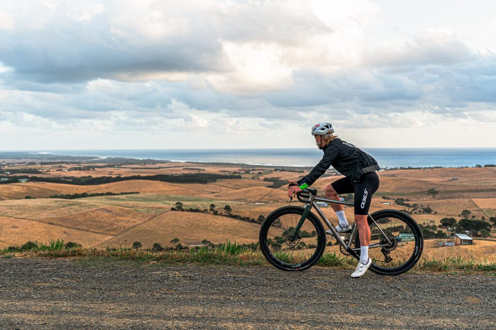 Rider on a Simpatico titanium gravel bike on an open gravel road with farmland and coast behind